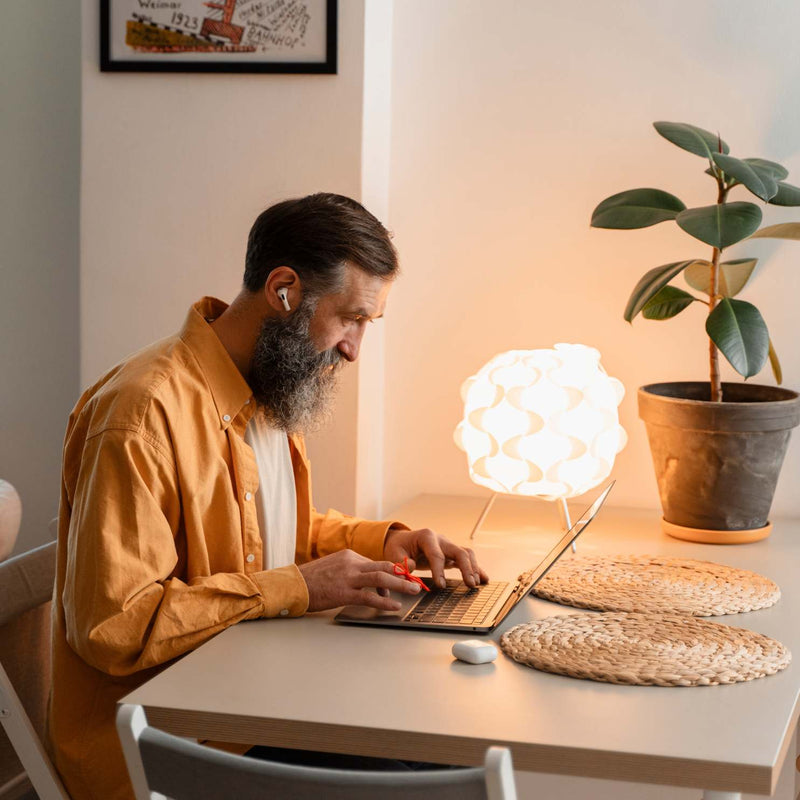 A man with a beard wearing a mustard yellow shirt sits at a desk using a laptop next to a glowing cloud-shaped lamp and a potted plant in a warm, cozy room.