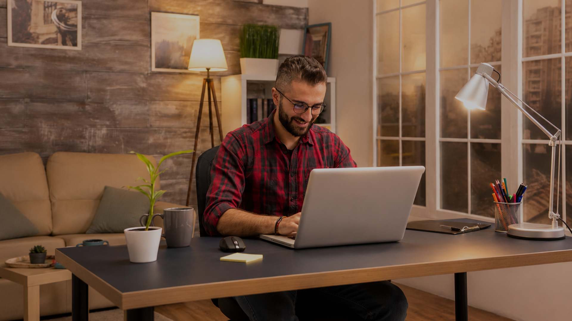 A man wearing glasses and a red plaid shirt sits at a wooden desk using a laptop in a cozy home office setting with warm lighting, a sofa, and a lamp.